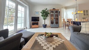 Un salon comprenant un canapé, une table basse avec un panier de fruits, un meuble pour la télévision, un buffet et un coin repas avec des chaises et une table.