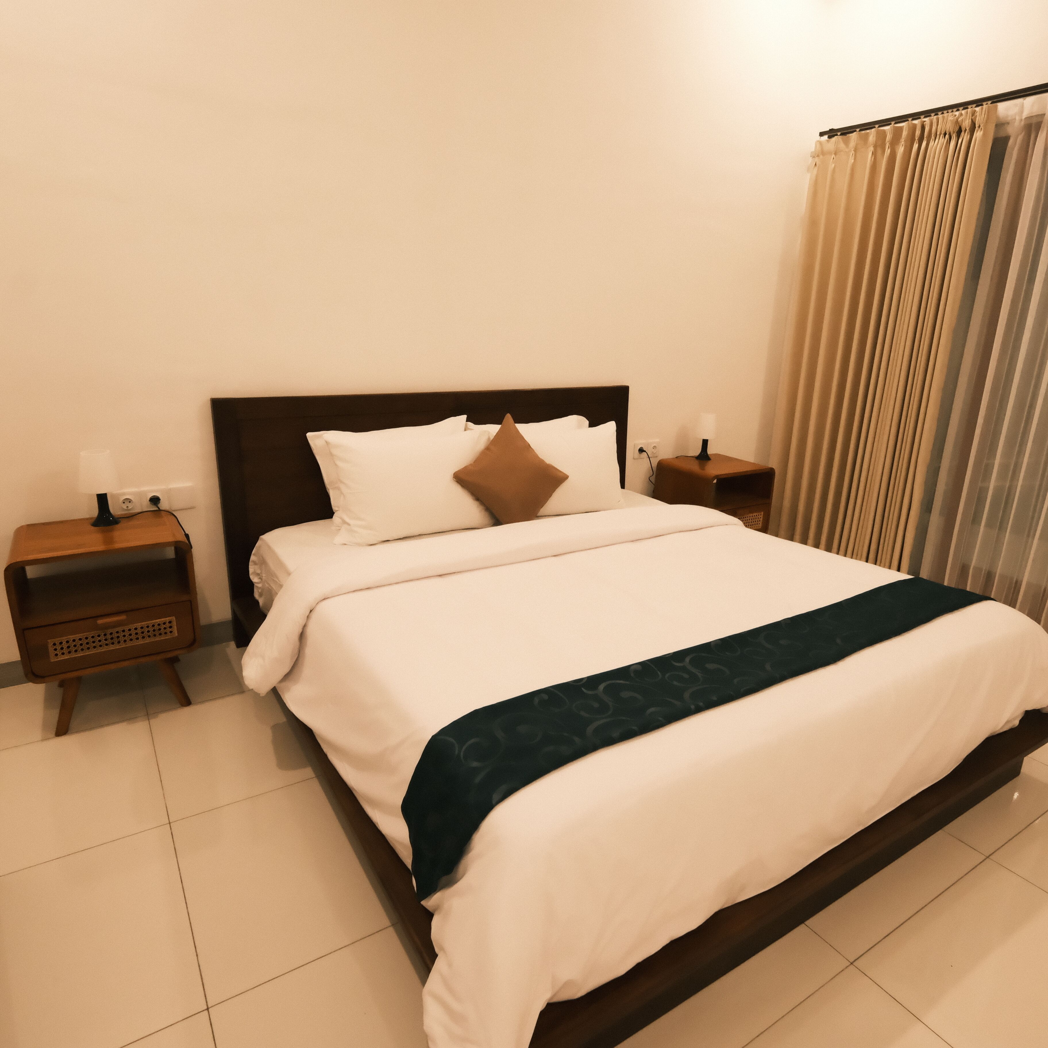 A neatly made bed with white and brown pillows, a dark headboard, and a dark blue bedspread. There are two wooden nightstands with lamps and a power outlet on the wall.