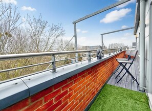Une terrasse sur le toit, dotée d’une rambarde en métal, d’un mur en briques et d’un petit espace aménagé avec une table et des chaises.