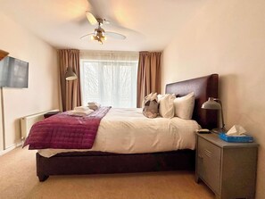 A hotel room with a bed, a nightstand, a lamp, a ceiling fan, a window with curtains, and a TV on the wall. - Split level unique large 2BDR apartment (Chelmsford)