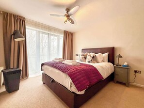 A hotel room with a bed, a ceiling fan, a nightstand with a lamp, and a view of the outdoors through large windows. - Split level unique large 2BDR apartment (Chelmsford)