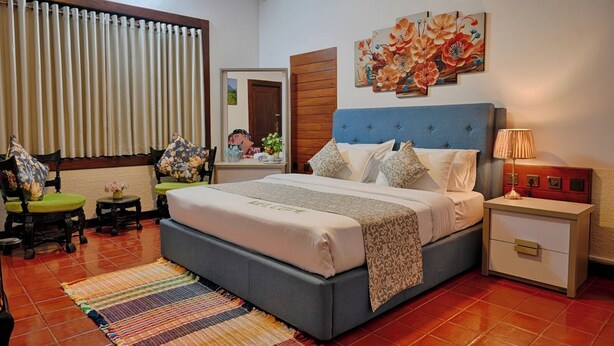 A bedroom with a large bed, a nightstand with a lamp, a chair with a floral cushion, a mirror, and a wall with a floral painting. - Cozy 2-bedroom apartment in serene Kandy with AC (Kandy)