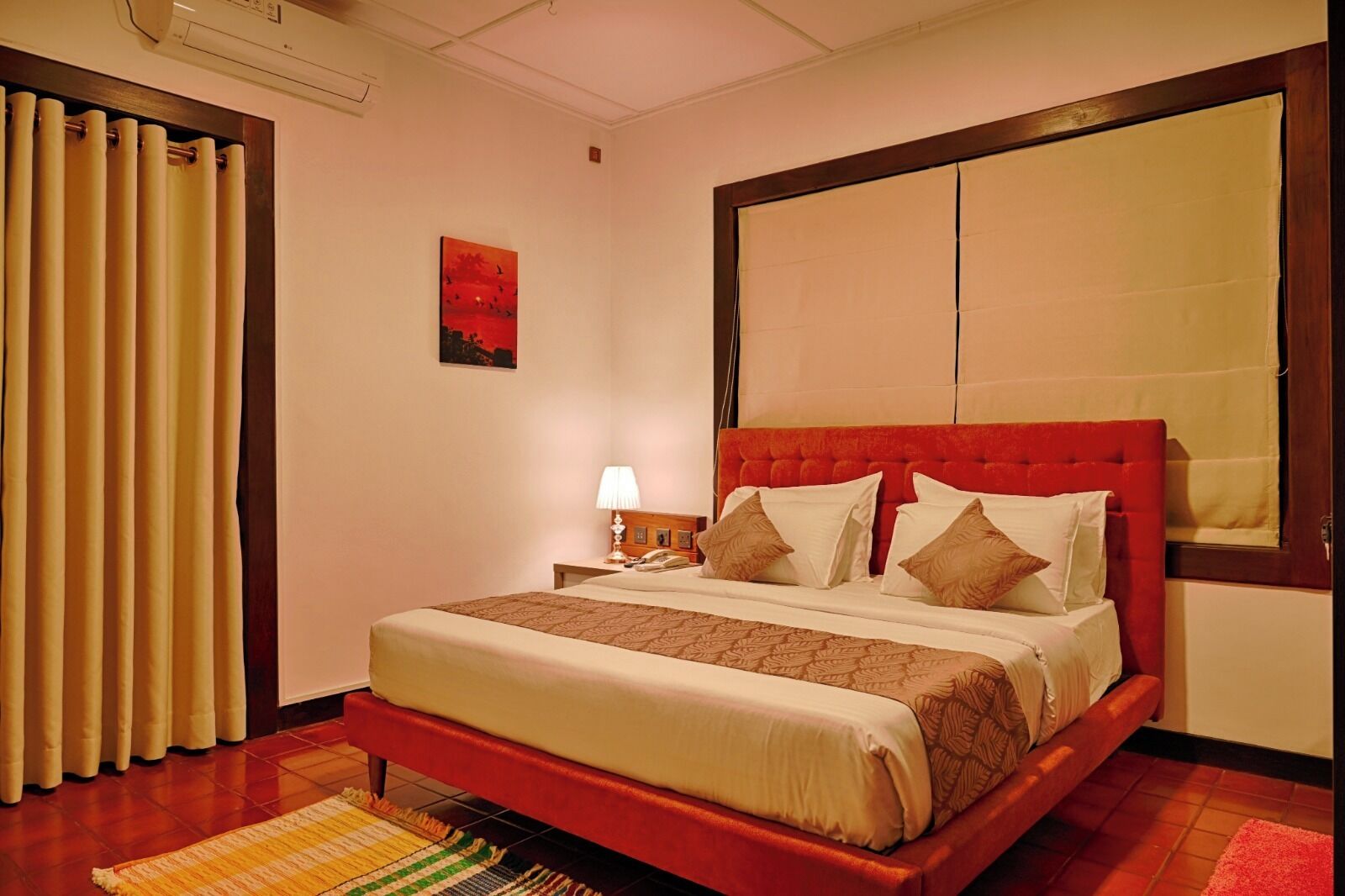 Habitación de hotel con una cama grande, una mesita de noche, una lámpara, un cuadro en la pared y una ventana con cortinas.
