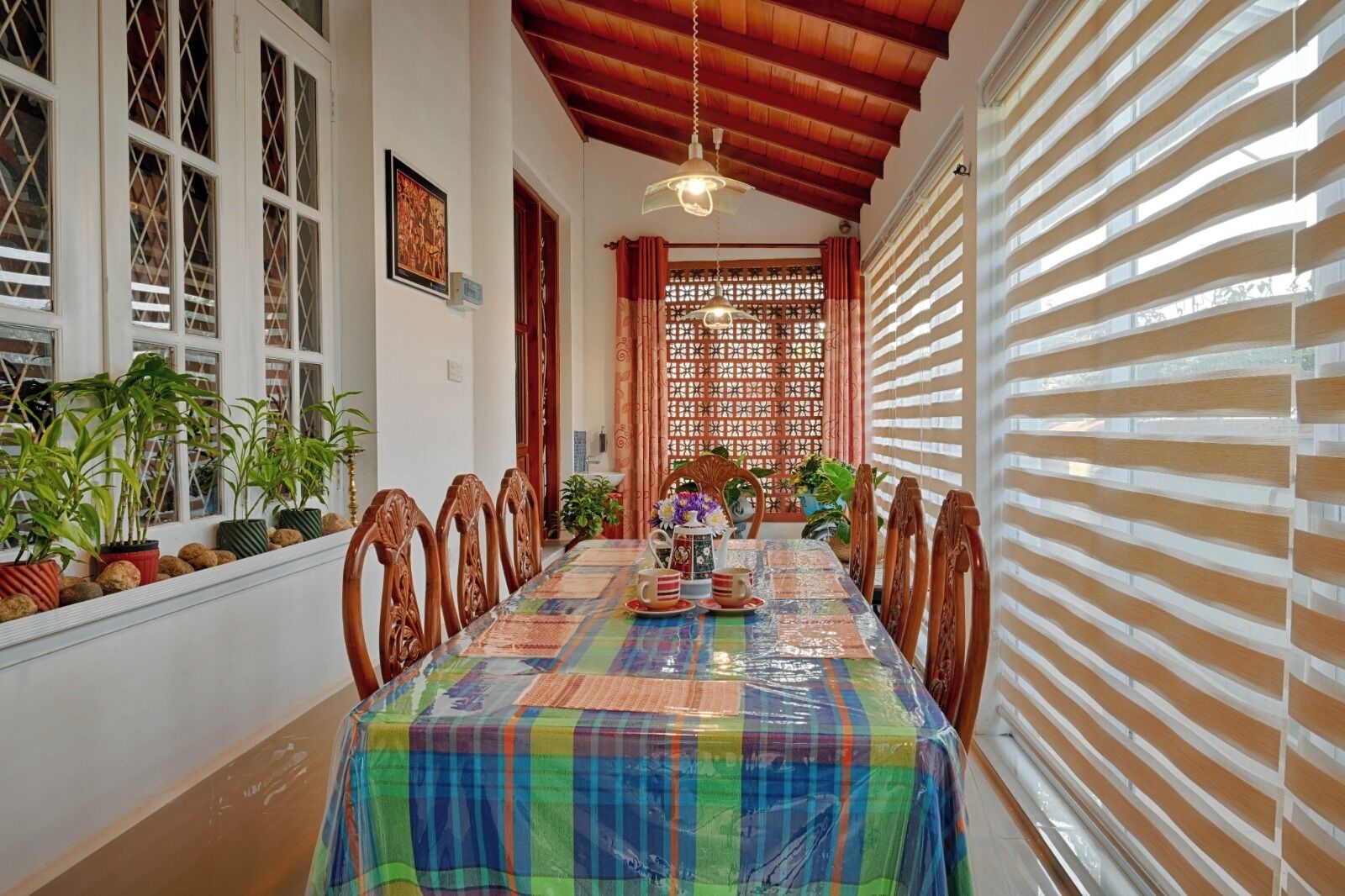 A dining area with a long table, chairs, potted plants, and a patterned tablecloth.