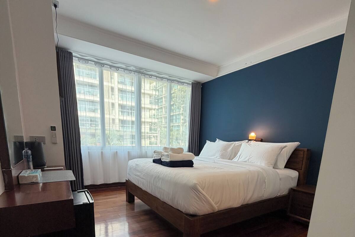 Una habitación de hotel moderna con suelo de madera, una cama grande con ropa blanca, una mesita de noche con lámpara y vistas a un edificio a través de ventanas amplias.