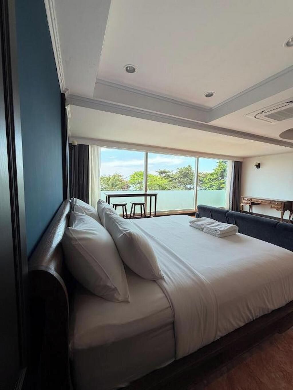 A large bed with white linens, a view of the outdoors through a large window, and a small table and chair set in the corner.
