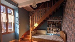 A bedroom with a wooden staircase, a bed, a bookshelf, and a window with lace curtains. - Typical stone house (Ribes)
