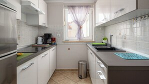 Eine kleine Küche mit weißen Schränken, einem Edelstahlkühlschrank, einem Fenster mit transparenten Vorhängen und einem Spülbecken mit einer grünen Fußmatte.