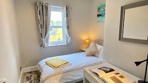 Une chambre à coucher comprenant un lit, un bureau, une chaise, une fenêtre avec des rideaux, une lampe et un tableau accroché au mur.