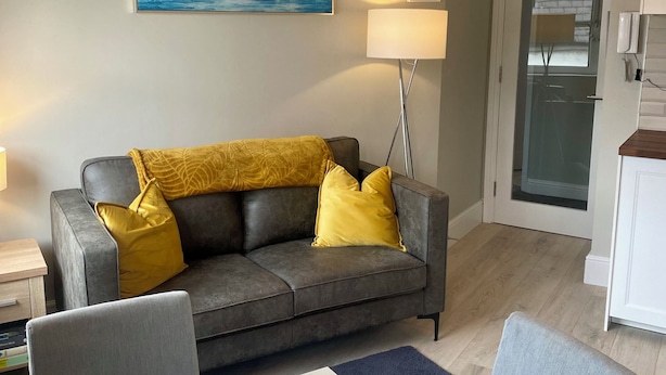 Un salon avec un canapé gris, des coussins jaunes, un tableau représentant un phare, une lampe et une porte en verre donnant sur une cuisine.