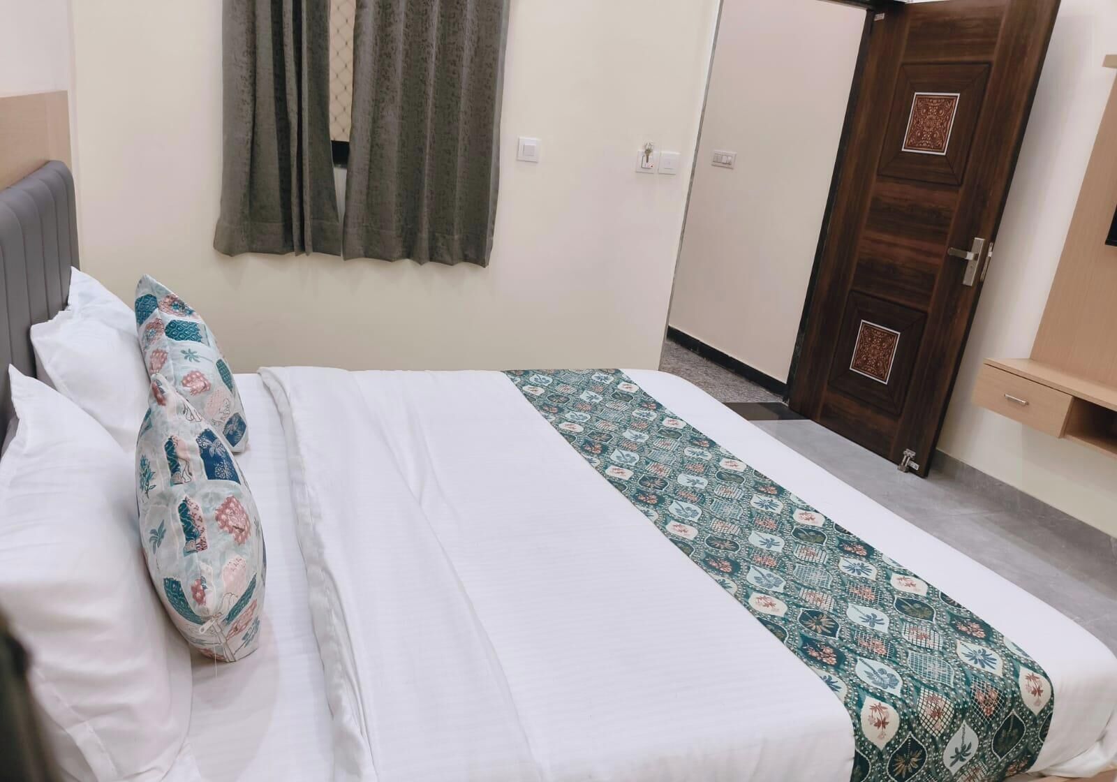 A neatly made bed with white linens and decorative pillows, a patterned bedspread, and a wooden wardrobe with a door.