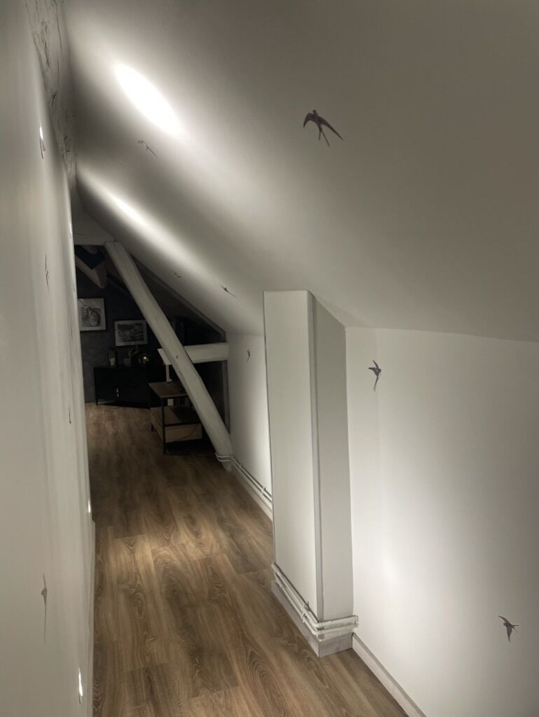 Un couloir étroit, doté d’un plancher en bois et de murs blancs, mène à un escalier et à une pièce où se trouve une grande armoire blanche.