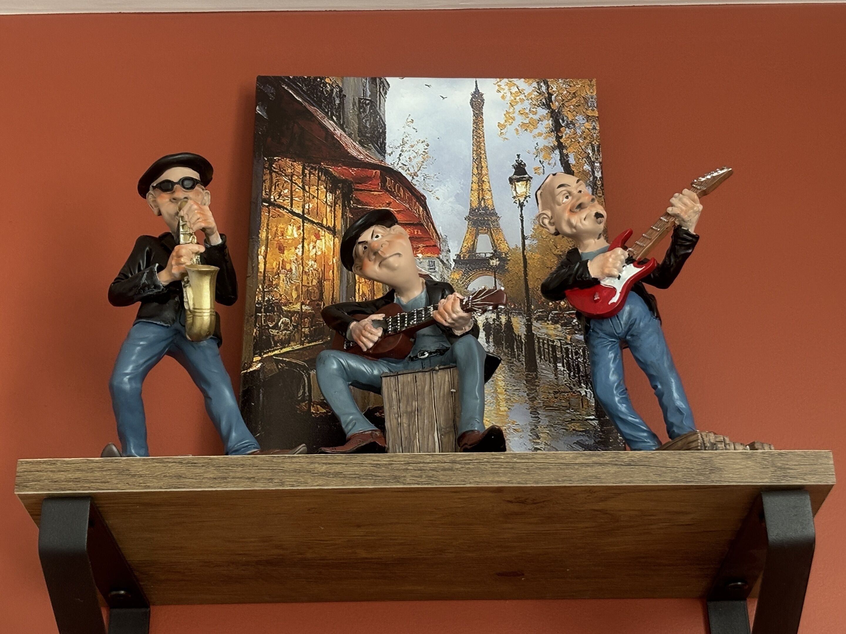 Three figurines of musicians on a shelf against a wall with a painting of Parisian street.