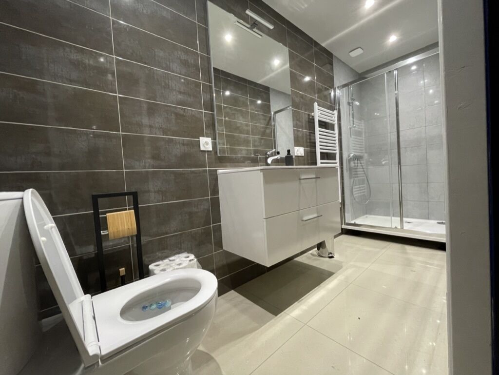 Un baño moderno con inodoro, lavamanos y zona de ducha, con baldosas oscuras y mampara de vidrio.