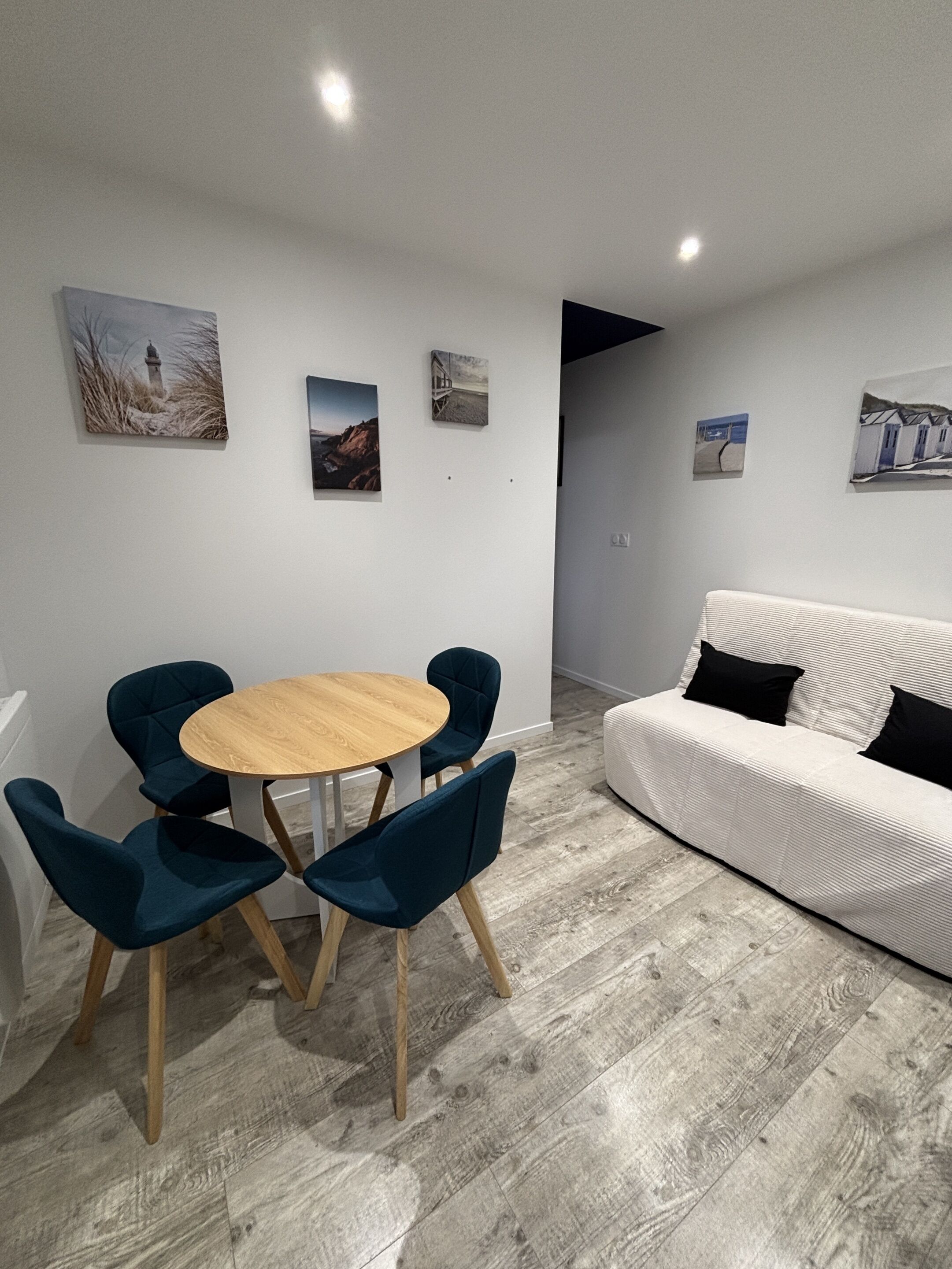 Een moderne woonkamer met een ronde houten tafel en vier donkere stoelen, een witte bank met zwarte kussens, en diverse ingelijste kunstwerken aan de muur.