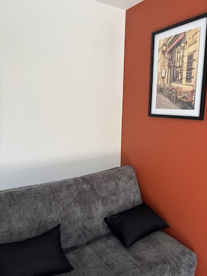 Un canapé gris avec des coussins noirs, un tableau encadré représentant une scène de rue et un mur blanc uni.