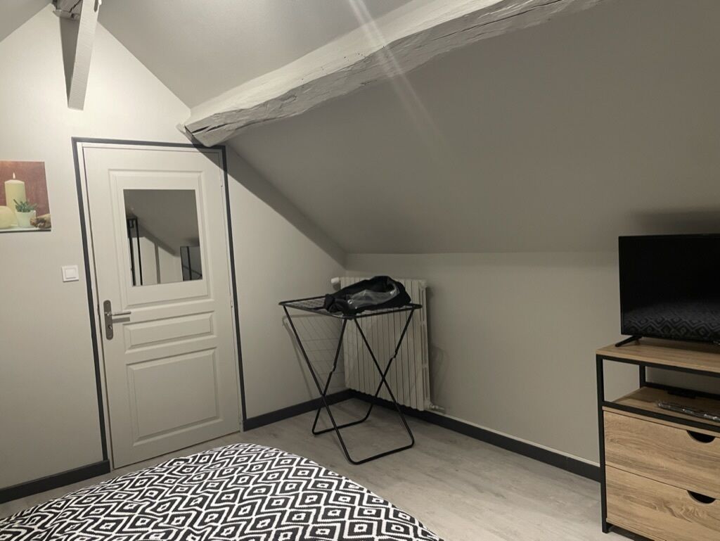 Une petite pièce bien éclairée, avec un plafond en pente, une porte blanche, un radiateur, un étendoir noir, une commode en bois et un tapis à motifs.