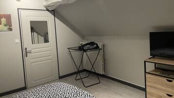 Une petite pièce bien éclairée, avec un plafond en pente, une porte blanche, un radiateur, un étendoir noir, une commode en bois et un tapis à motifs.