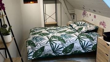 Un dormitorio con una cama, una escalera de madera, un tapete tejido, un estante con plantas y un cuadro de un atardecer.