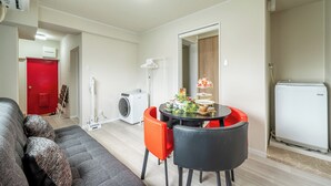 Ein modernes Wohnzimmer mit grauem Sofa, einem Esstisch mit Stühlen, einer Waschmaschine und einem kompakten Kühlschrank.