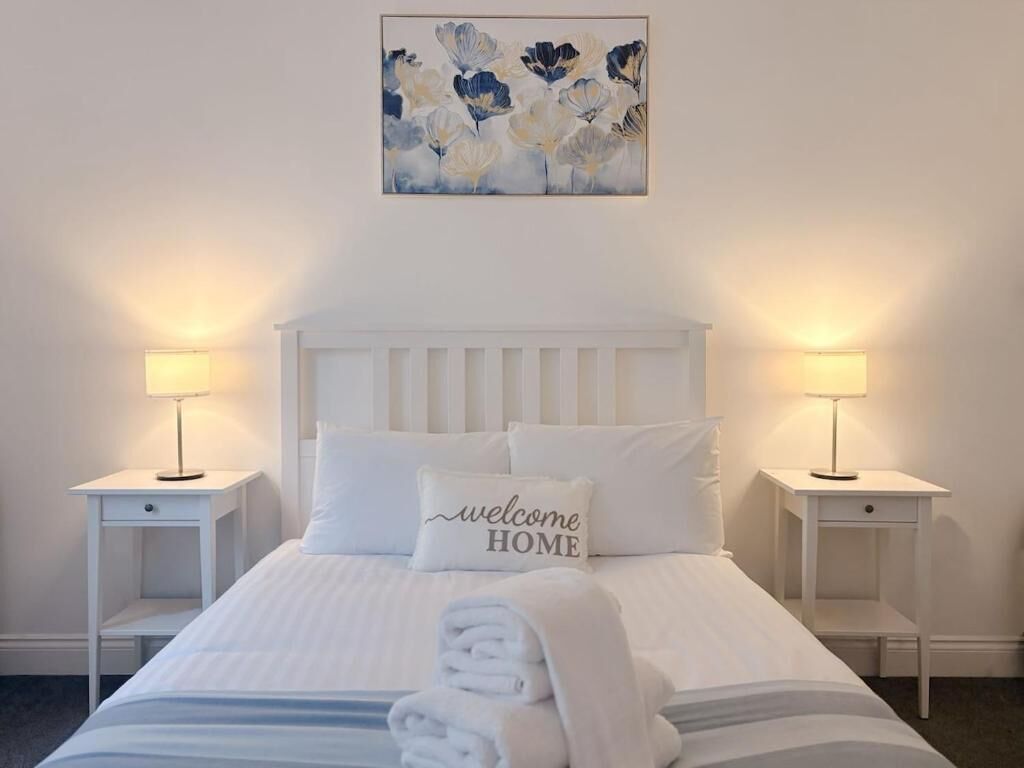 A neatly made bed with a 'welcome home' pillow, two bedside lamps, and a floral painting on the wall.