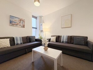 Ein Wohnzimmer mit zwei dunklen Sofas, einem weißen Couchtisch und einer Blumenvase. An den Wänden hängen zwei gerahmte Bilder.