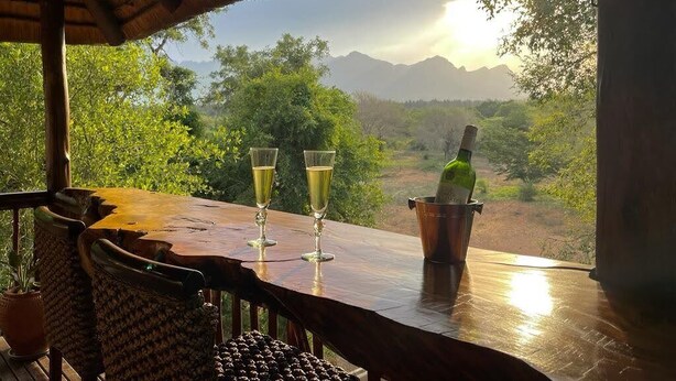 Une table en bois, avec deux coupes de champagne et une bouteille dans un seau à glace, donnant sur un paysage luxuriant et montagneux.