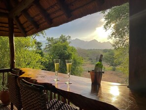 A wooden table with two glasses of champagne and a bottle in a bucket, overlooking a lush landscape with mountains. - Master Bedroom (houedspruit)