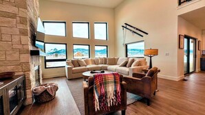 A spacious living room with a stone fireplace, a large sectional sofa, and a view of mountains through multiple windows. - Modern Family Retreat w/ Hot Tub, Teton Views, + A/C (Driggs)