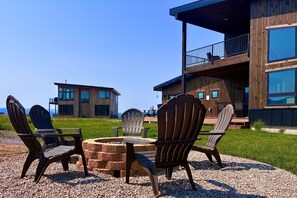 Un espacio al aire libre moderno con sillas Adirondack y un hogar para fuego, con vistas a un edificio de dos pisos con ventanas amplias y un balcón.