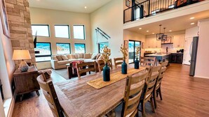 A spacious living room with a wooden dining table, comfortable seating, and large windows offering a view of the outdoors. - Modern Family Retreat w/ Hot Tub, Teton Views, + A/C (Driggs)