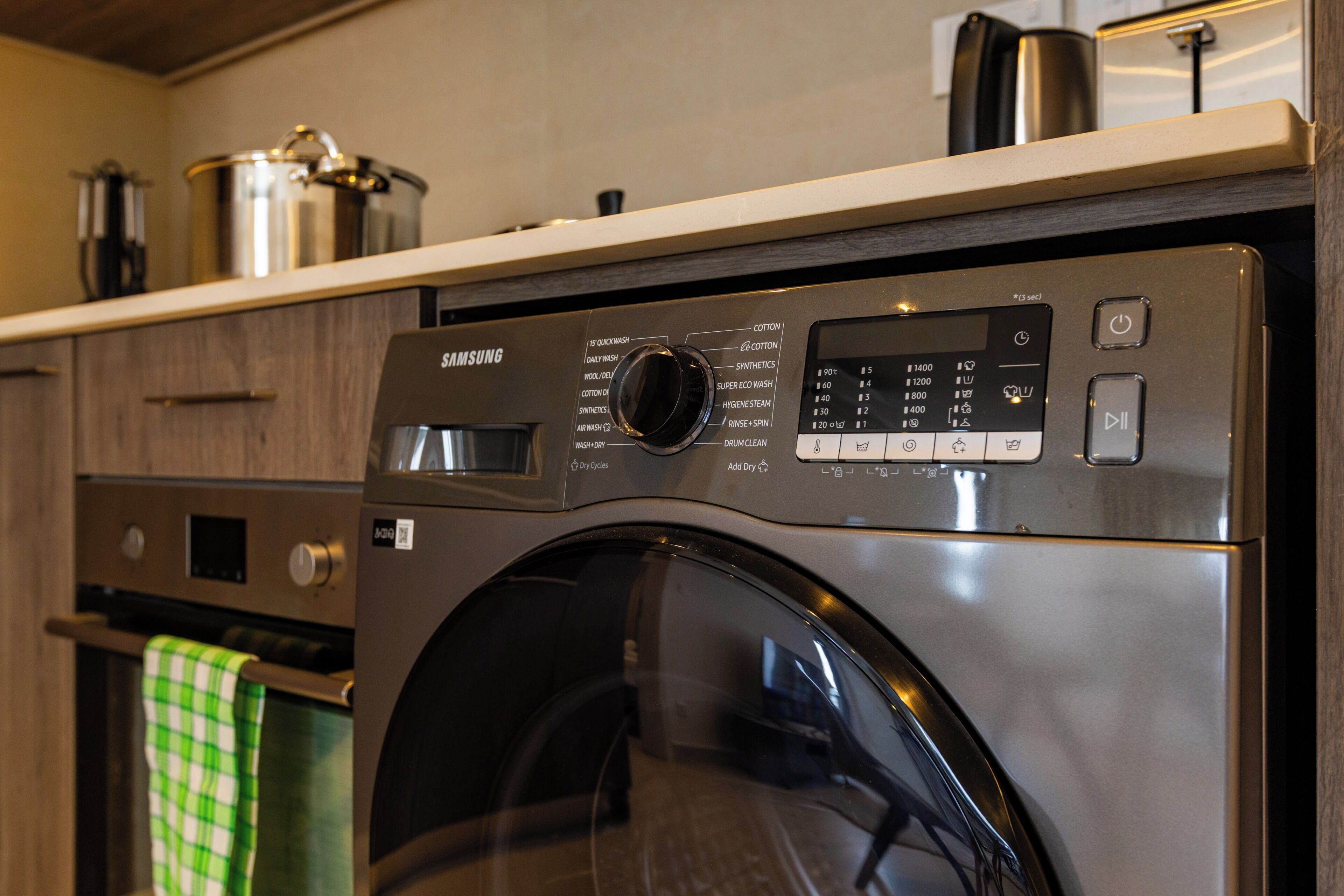 A Samsung front-loading washing machine with a control panel, a stainless steel pot, and a green checkered towel on a wooden countertop.