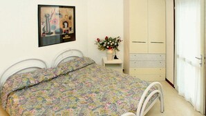 A bedroom with a bed, a wardrobe, a small table with a vase, and a picture on the wall. - Lignano Riviera apartment with pool access (Lignano Sabbiadoro)