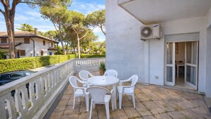 Eine Terrasse mit rundem Tisch und Stühlen, eine weiße Klimaanlage und Blick auf die Umgebung mit Bäumen und anderen Gebäuden.