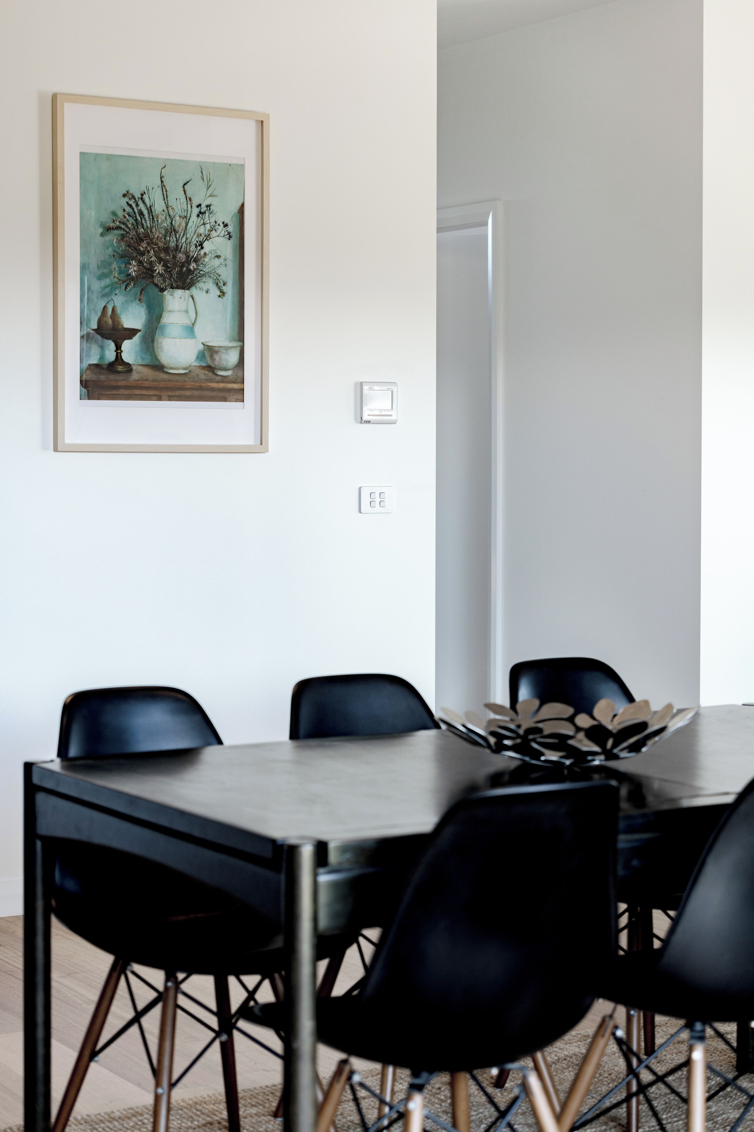 Een eethoek met een tafel en stoelen, een ingelijst schilderij aan de muur en een deur op de achtergrond.