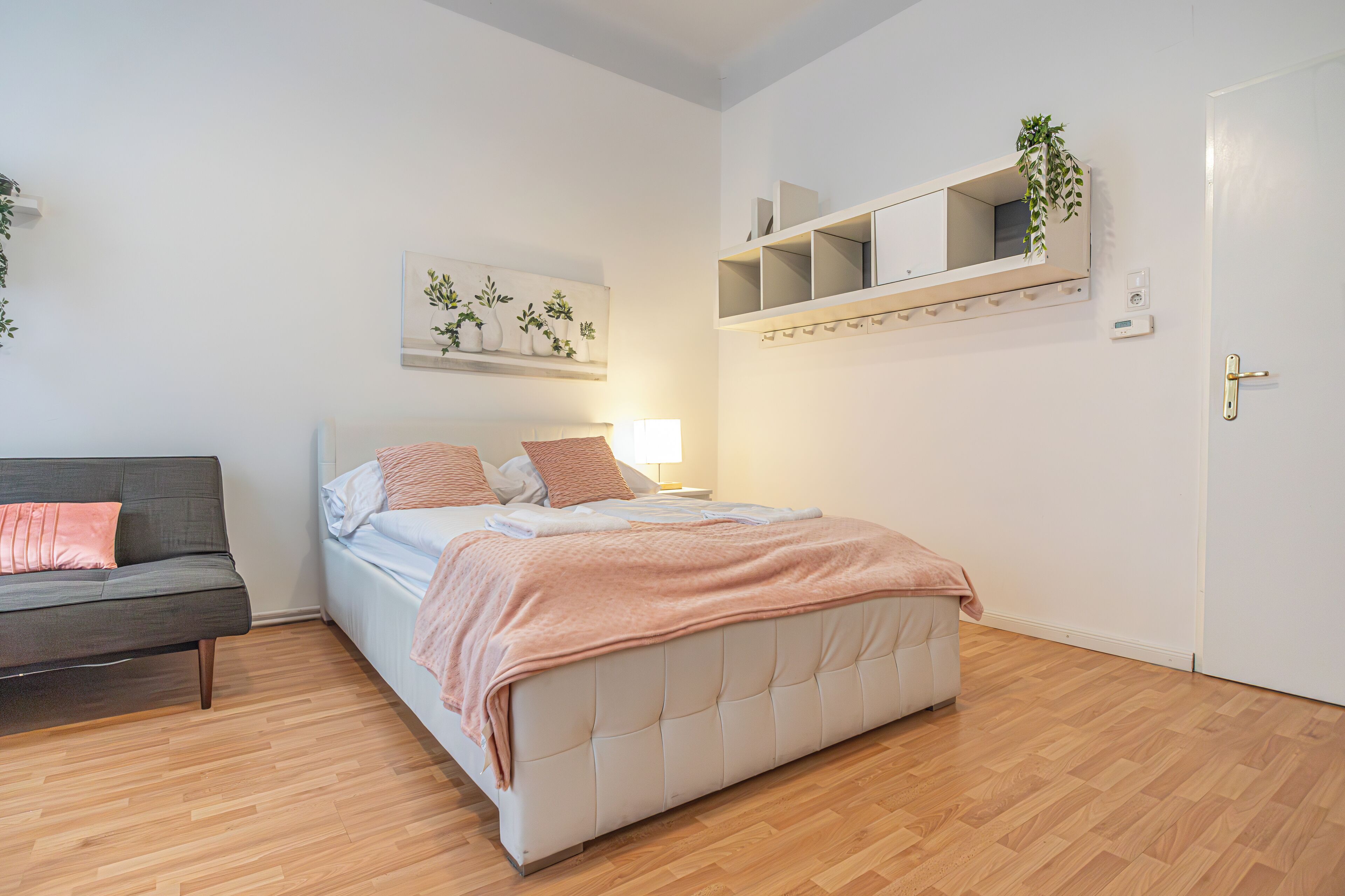 A bedroom with a bed, a sofa, a bedside table, a wall-mounted shelf with a plant, and a door.