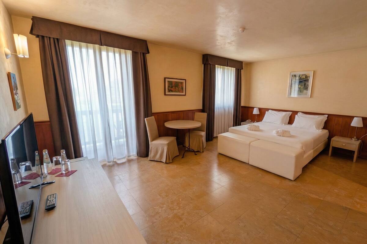 A hotel room with a bed, a desk with a TV, a chair, a small table, and a window with curtains.