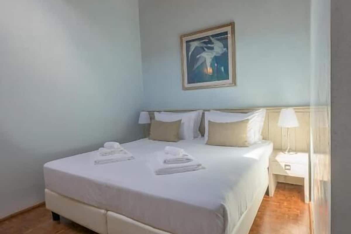 Quarto com teto de madeira, cama com roupa de cama branca, mesa de cabeceira e um quadro na parede.