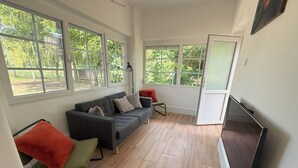 A modern living room with a grey sofa, green and orange chairs, a flat-screen TV, and large windows offering a view of greenery.