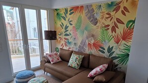 Ein Wohnzimmer mit braunem Sofa, gemusterten Kissen, einem runden Tisch und lebhaftem tropischem Blattenteppich an der Wand.