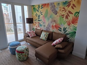 Un salon avec un canapé marron, des coussins à motifs, une table ronde et un papier peint aux motifs de feuilles tropicales aux couleurs vives.