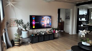 Een woonkamer met een flatscreen televisie aan de muur, houten vloeren en diverse potplanten op een zwarte console tafel.