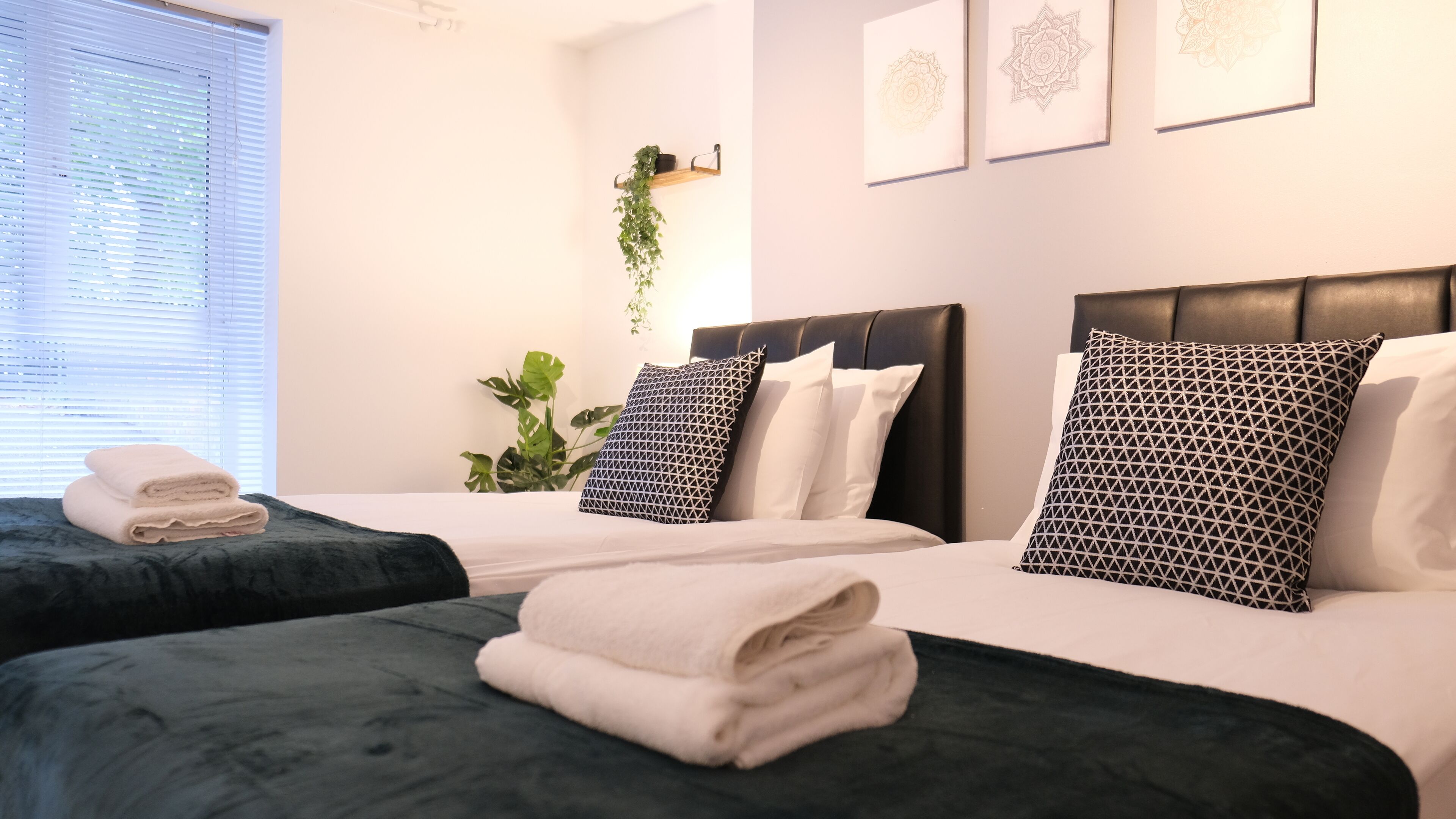 Ein Hotelzimmer mit zwei Betten, jeweils mit dunkelgrüner Bettdecke und weißen Kissen. An der Wand befinden sich gerahmte Kunstwerke, ein Blumentopf und ein Fenster mit Jalousien.