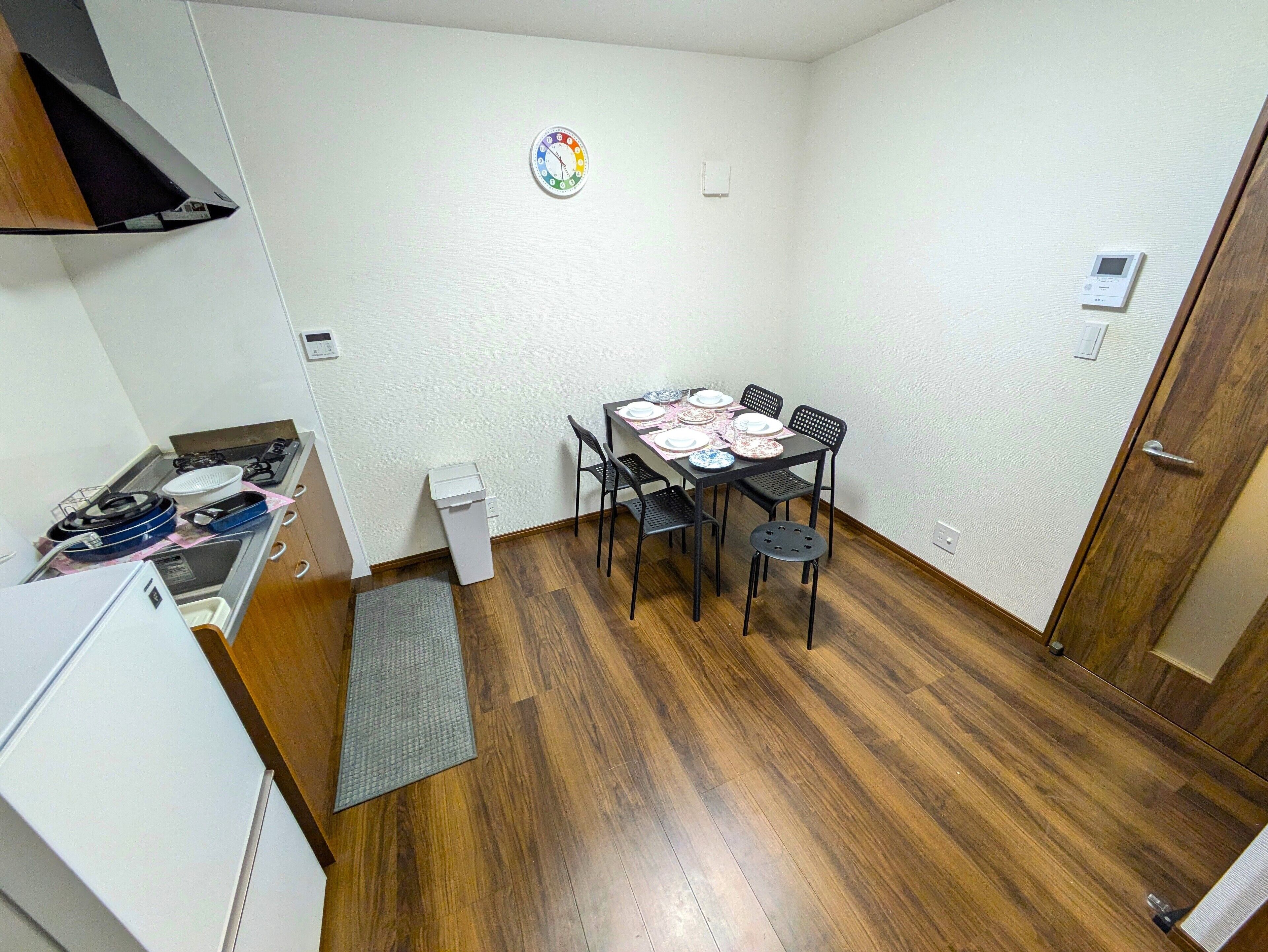 Une petite cuisine compacte, équipée d’une cuisinière, d’un évier et d’une petite poubelle. On y trouve une table à manger avec des chaises et de l’assiette déjà mise en place pour un repas. La pièce est dotée d’un plancher en bois et de murs blancs.