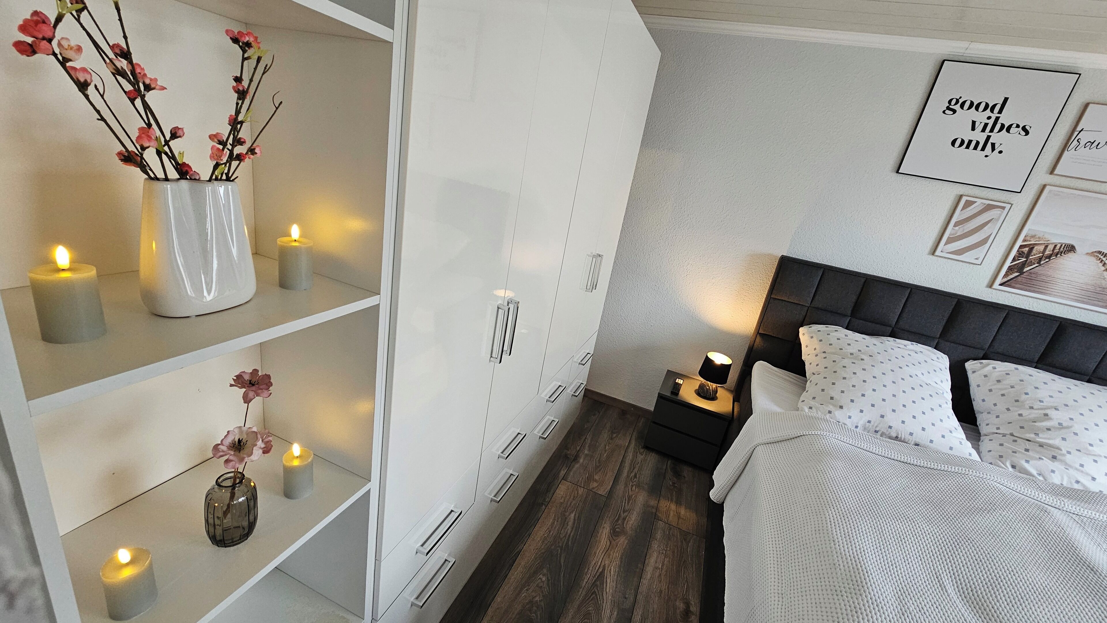 Un dormitorio con cama, mesitas de noche, armario y elementos decorativos como velas y flores.
