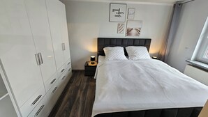 Une chambre dotée d’une grande armoire, d’un lit recouvert d’une couette blanche à texture particulière, de deux lampes de chevet et de décorations murales, dont un panneau affichant l’inscription « bonnes ondes seulement ».