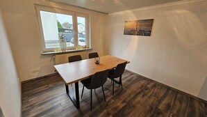 Une salle de conférence avec une table en bois, quatre chaises, un radiateur, une fenêtre donnant sur une maison et un tableau représentant un paysage urbain accroché au mur.