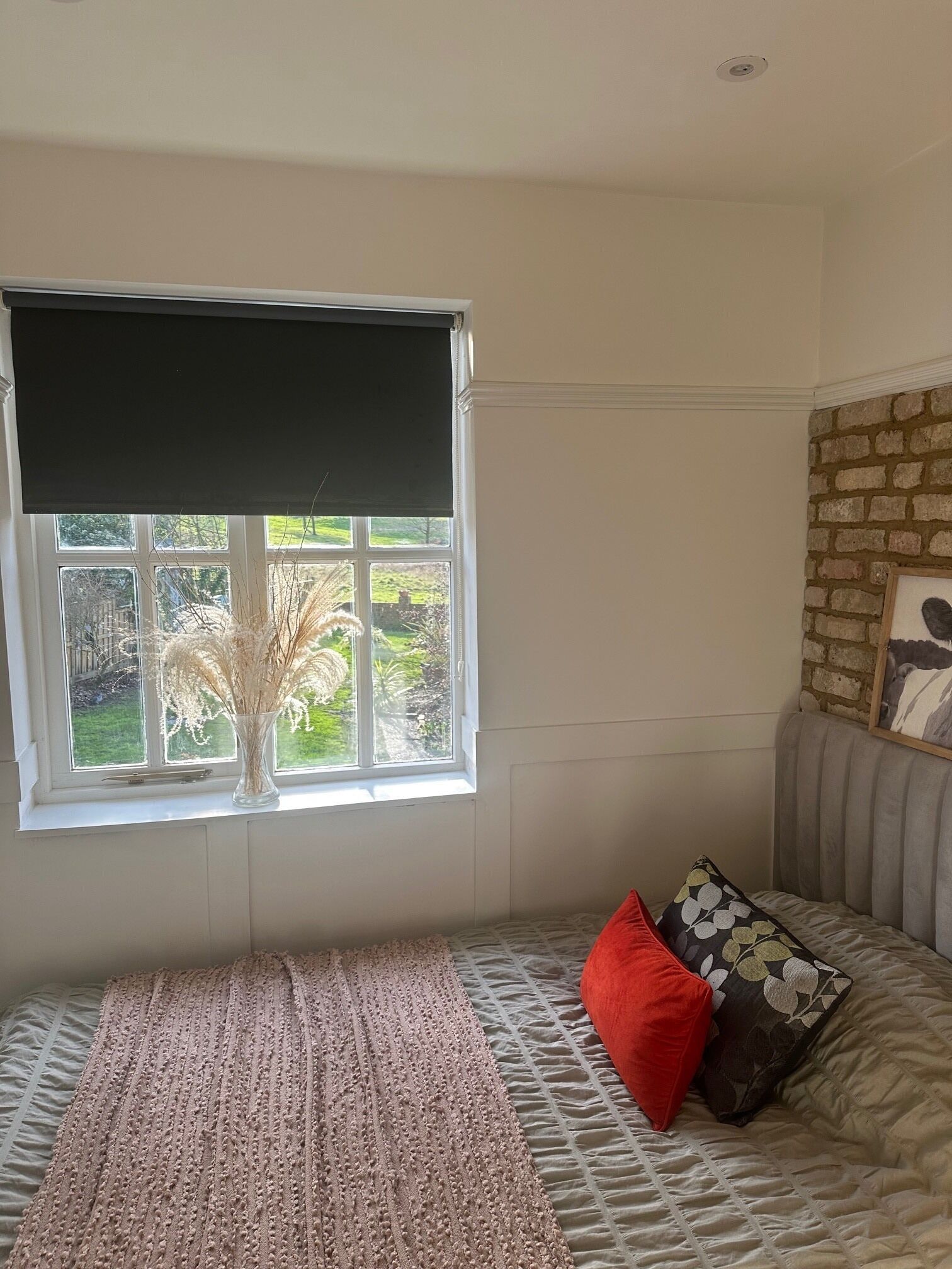 A room with a window, a bed with patterned bedding, a red pillow, and a framed picture on the wall.