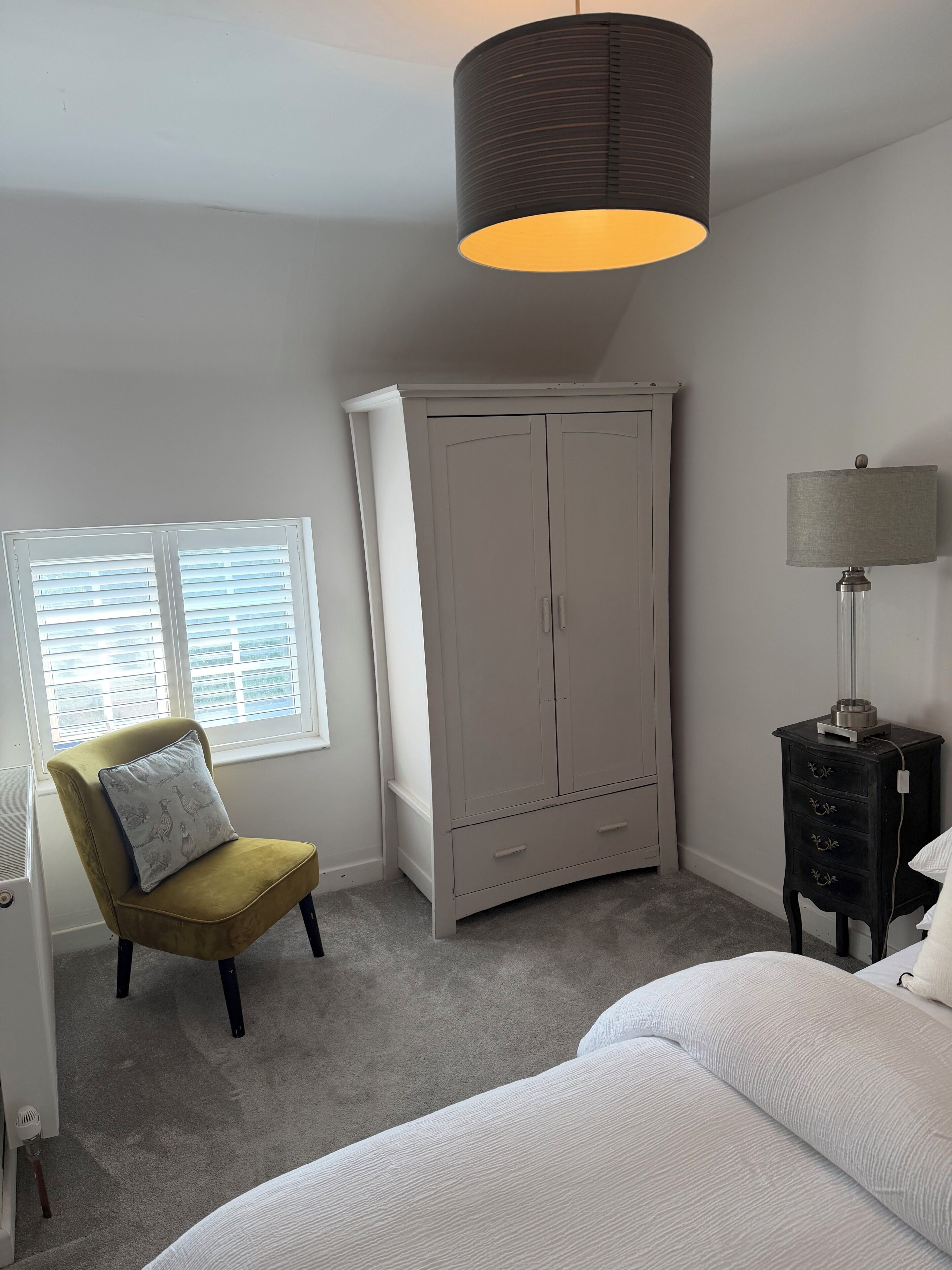 A bedroom with a bed, a chair, a wardrobe, a lamp, and a window with blinds.
