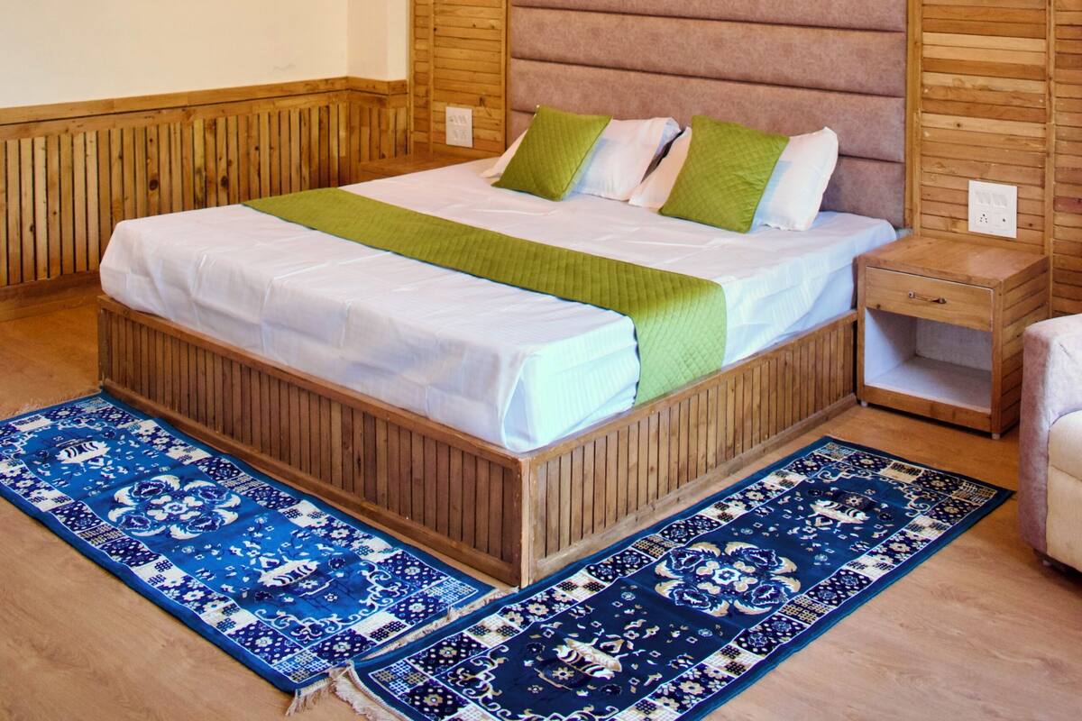 A bedroom with a wooden ceiling, a bed with a green and white bedspread, a blue patterned rug, and a nightstand.